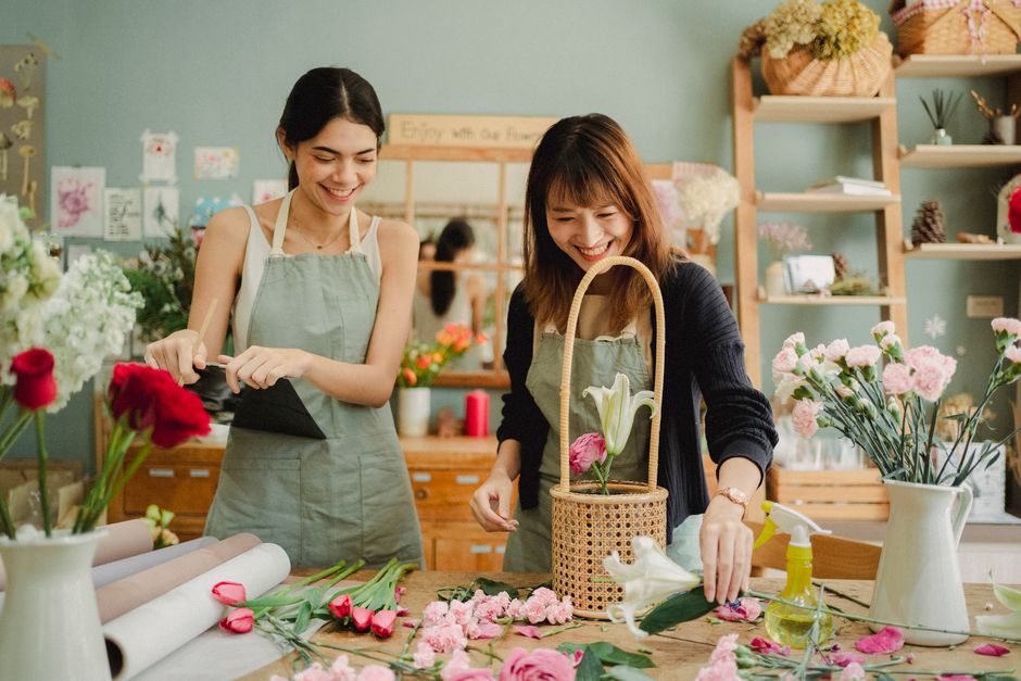 Smiling multiracial florists in aprons standing near wooden table and preparing bouquets for order in cozy floristry shop in daylight