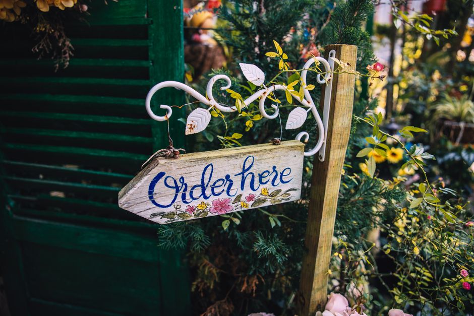 A rustic 'Order Here' sign decorates a vibrant garden with colorful flowers.