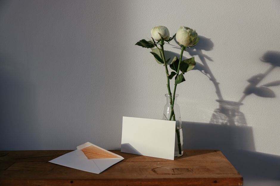 A minimalist scene showcasing white roses in a vase next to a blank card and envelope on a wooden surface.