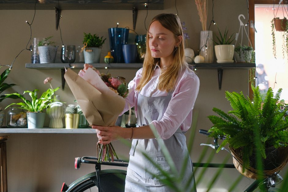 A female florist arranges a fresh bouquet in a cozy indoor setting with various plants and decor.