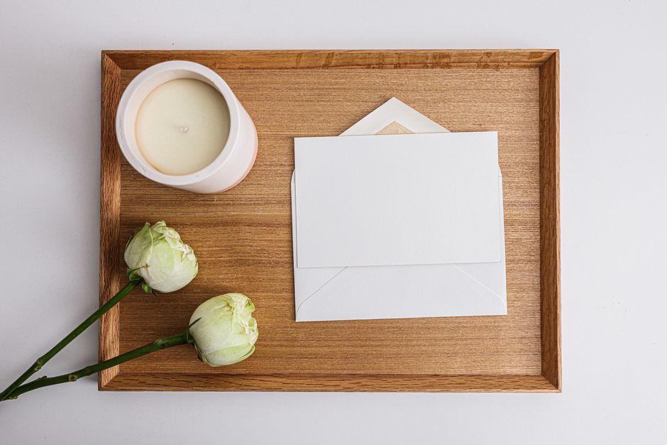 Flat lay of white envelope, flowers, and candle on a wooden tray, perfect for invitations.
